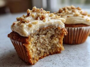 Close-up of a banana walnut cupcake with a bite taken out, showing the moist crumb and cream cheese frosting topped with chopped walnuts.