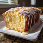 A loaf of moist cream cheese pound cake drizzled heavily with a thick white glaze, sitting on a white rectangular plate.