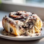 A close-up shot of a single, gooey oreo cinnamon rolls covered in thick white icing and cookie crumbles.