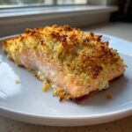 Close-up of a flaky salmon fillet with a golden, crispy parmesan crusted baked salmon topping on a white plate.