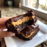 A hand holds up one half of a rich, fudgy peanut butter stuffed brownie, revealing the thick peanut butter filling.