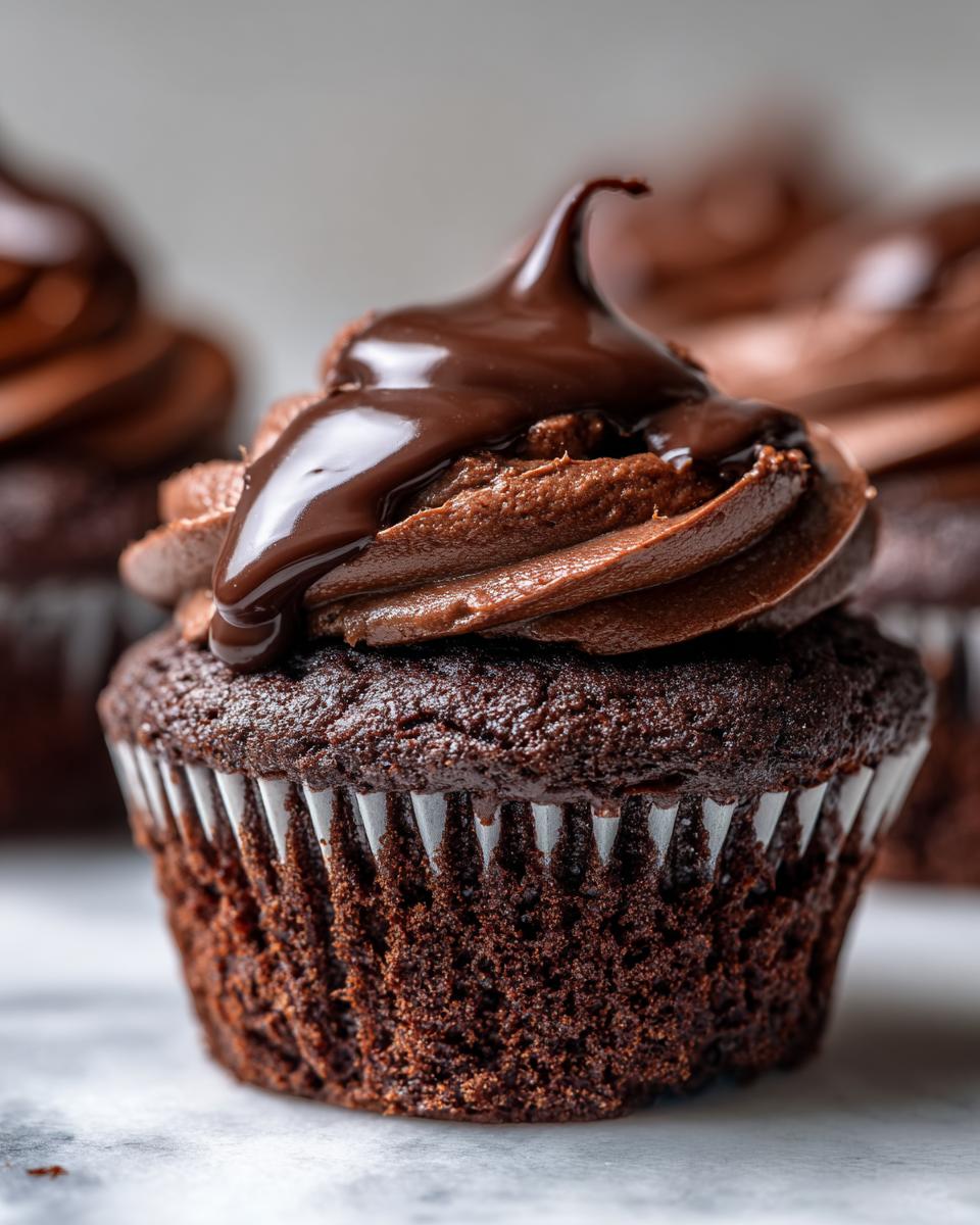 Close-up of a decadent chocolate cupcake topped with chocolate frosting and a rich chocolate ganache drizzle, part of a batch of raspberry chocolate cupcakes.