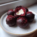 A stack of chocolate-dipped red velvet cake balls, one cut open showing the white cream cheese filling.