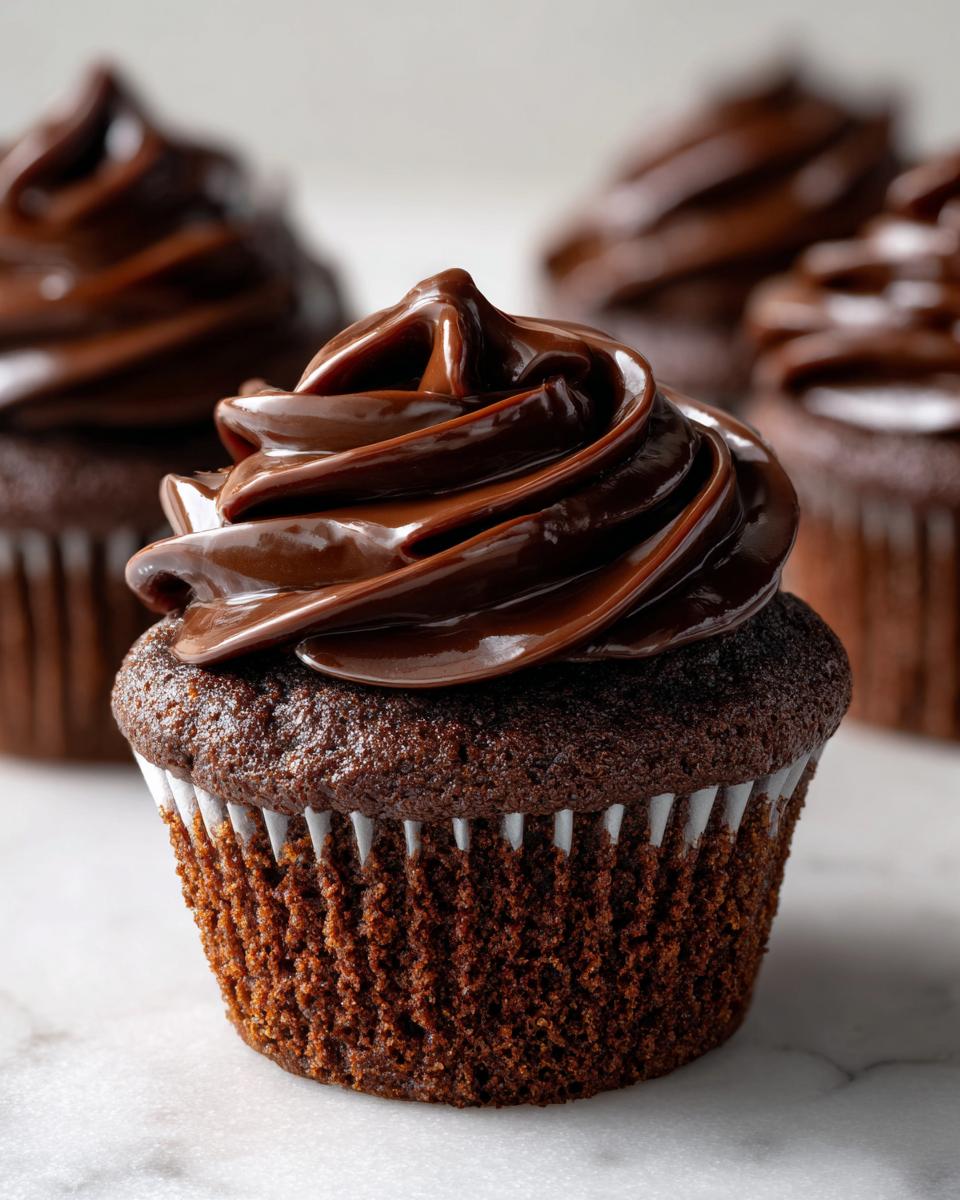 Close-up of a decadent chocolate cupcake with glossy, swirled chocolate frosting, part of a batch of raspberry chocolate cupcakes.