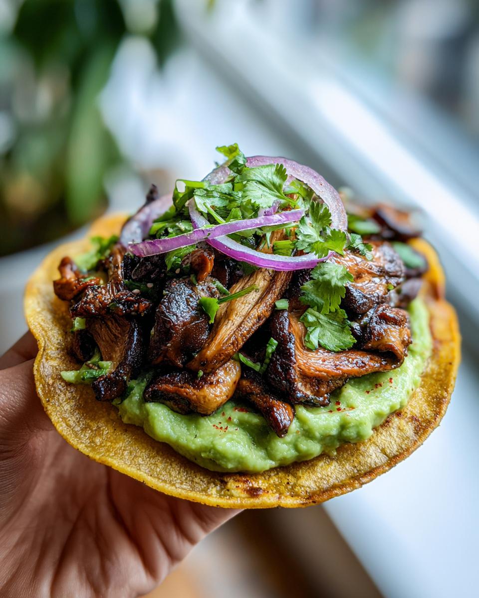 A hand holding a single roasted mushroom tostadas topped with creamy avocado, dark roasted mushrooms, red onion, and cilantro.