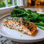 A close-up of a flaky, herb-crusted salmon fillet served alongside bright green broccolini for a quick salmon broccolini dinner.