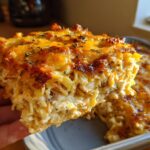 A hand lifts a cheesy, baked slice of Monterey chicken spaghetti casserole topped with melted cheddar and herbs.