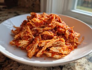 A close-up of shredded crockpot italian chicken mixed generously with a rich, red tomato sauce, served on a white plate.