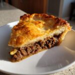 Close-up of a thick slice of homemade meat pie showing rich, seasoned ground filling and a golden, flaky pastry crust.