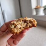 A hand holding a half-eaten, soft cinnamon toast crunch cookie, showing the oat-filled, chewy interior.
