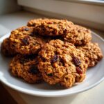 A close-up of several soft oatmeal raisin cookies piled on a white plate, catching sunlight.