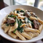 Close-up of creamy steak spinach alfredo penne skillet served in a white bowl, featuring penne pasta, beef chunks, and wilted spinach.