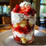 Layered strawberry shortcake parfaits in a mason jar with cake, strawberries, and whipped cream.