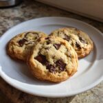 Three freshly baked classic chocolate chip cookies with melted dark chocolate chunks resting on a white plate.