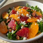 Close-up of a vibrant citrus salad featuring slices of blood orange and navel orange over arugula, topped with feta cheese and pecans.