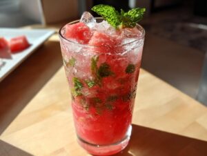 A tall glass filled with a vibrant red watermelon mint mojito, crushed ice, mint leaves, and watermelon chunks.
