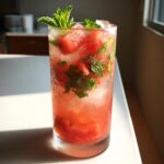 A tall, sweating glass filled with a refreshing watermelon mint mojito, garnished with fresh mint sprigs and watermelon chunks.