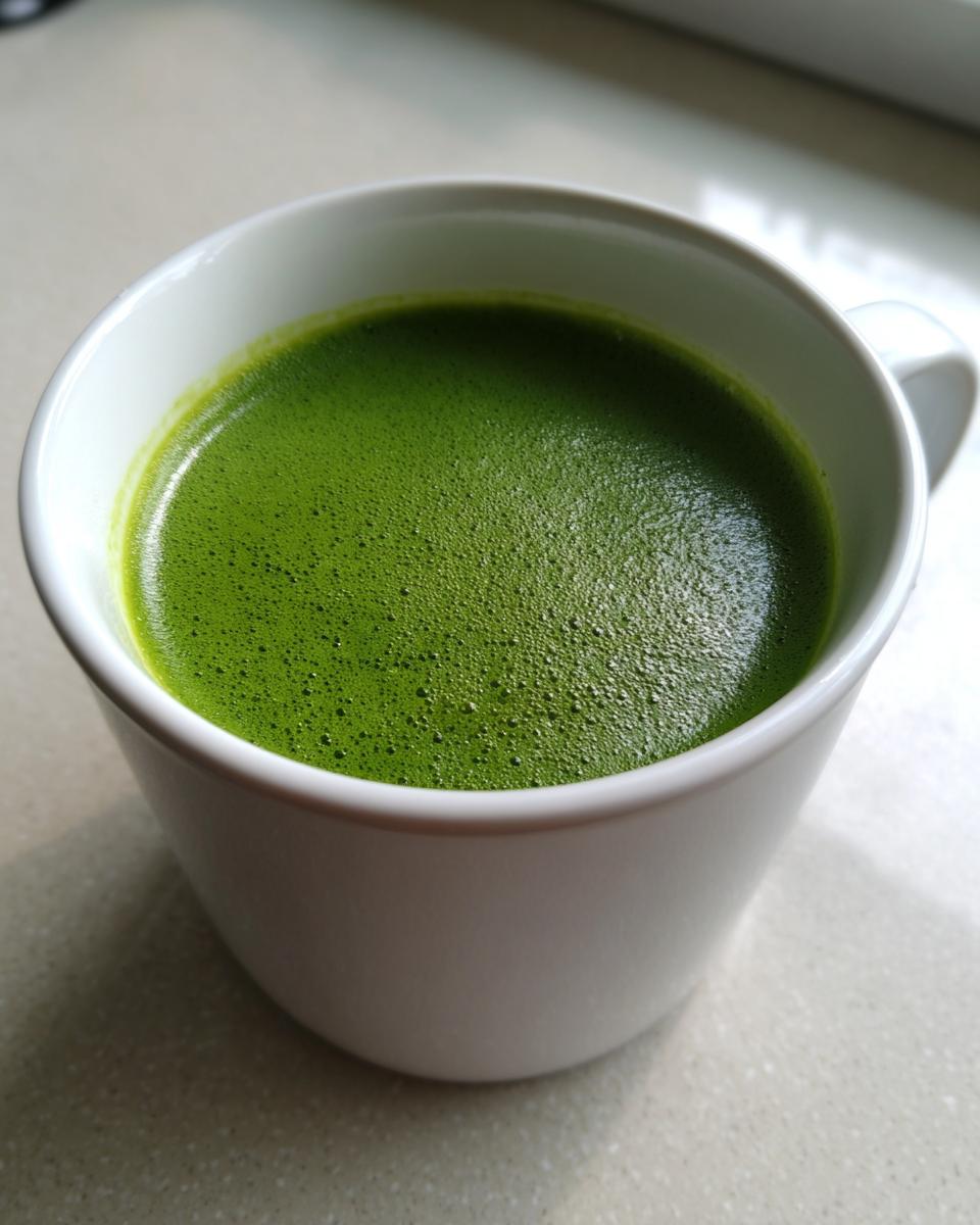 Close-up of a bright green, foamy liquid resembling Creamy Asparagus Soup served in a white mug.