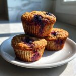 Three golden brown High Fiber Blueberry Bran Muffins stacked on a white plate, showing juicy blueberry bursts.