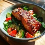 Close-up of a perfectly cooked Honey Glazed Salmon Bowl featuring a shiny, sesame-topped salmon fillet over quinoa, spinach, cucumber, and tomatoes.