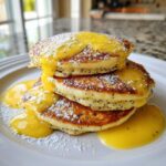A close-up stack of three fluffy Lemon Poppy Seed Pancakes drizzled with bright yellow lemon curd and dusted with powdered sugar.