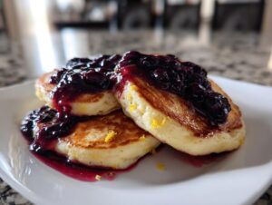 Three fluffy Lemon Ricotta Pancakes stacked and topped with rich blueberry sauce and lemon zest.