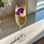 A flute of sparkling wine featuring edible purple pansies as part of a Mimosa Bar with Floral Ice Cubes.