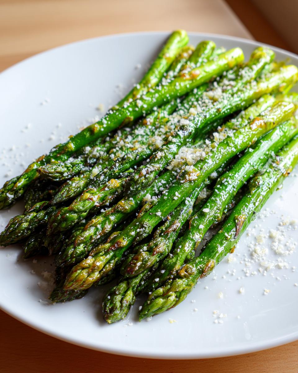A close-up of bright green Roasted Asparagus with Lemon and Parmesan sprinkled on top, served on a white plate.