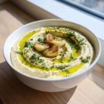 A white bowl filled with creamy Roasted Garlic Hummus, topped with roasted garlic cloves, olive oil, and fresh parsley.