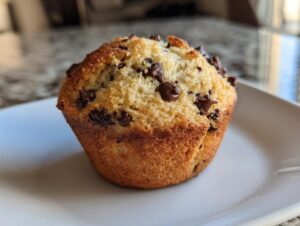 A close-up of a golden-brown Sourdough Discard Chocolate Chip Muffin studded with melted chocolate chips on a white plate.