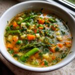 A close-up, appetizing view of a white bowl filled with vibrant Spring Vegetable Minestrone soup, featuring carrots, peas, asparagus, and herbs.